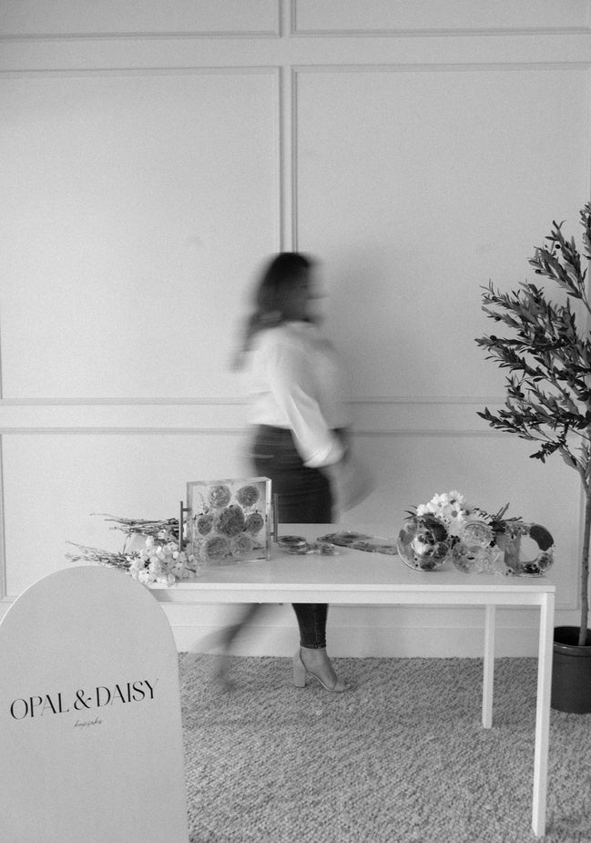 Person walking past a table with floral arrangements and 'Opal & Daisy' branding in a room.